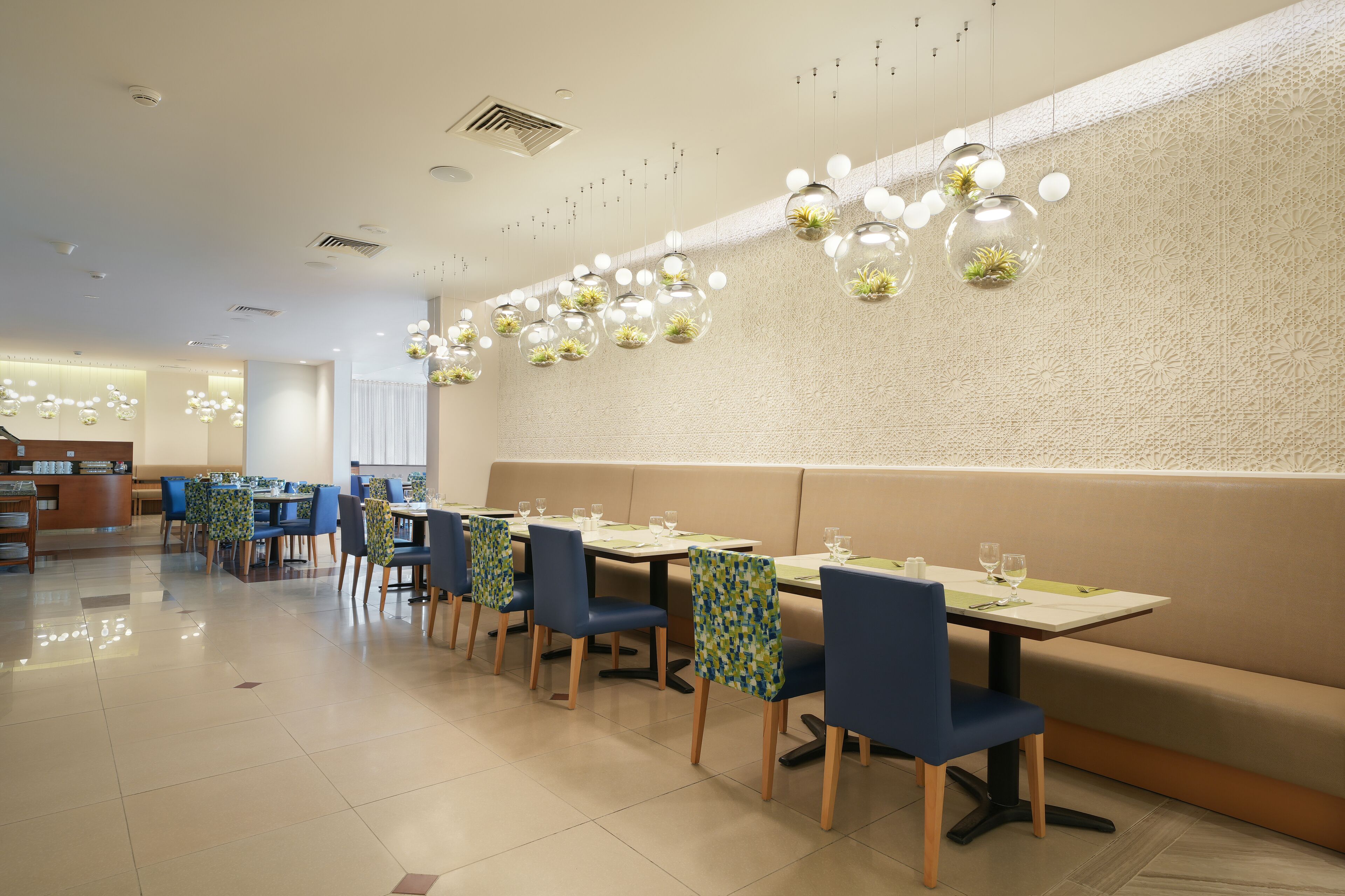 A modern dining area with booths and tables, featuring a patterned wall and hanging spherical light fixtures with plants.