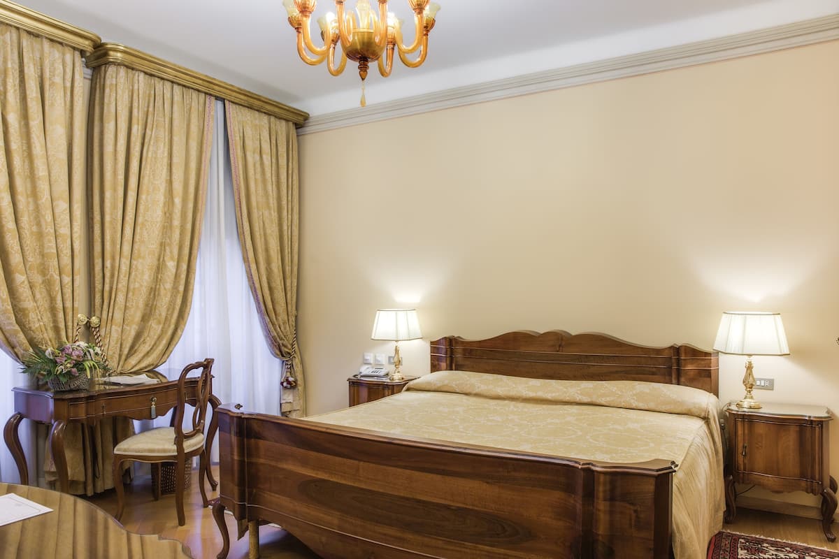 A hotel room with a wooden bed, two bedside tables with lamps, a desk with a chair, and a large window with curtains.