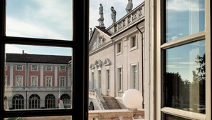A view through a window of a building with classical architecture, including a balcony with chairs and a table, and a statue on the roof. - Villa Fenaroli Palace Hotel (Rezzato)