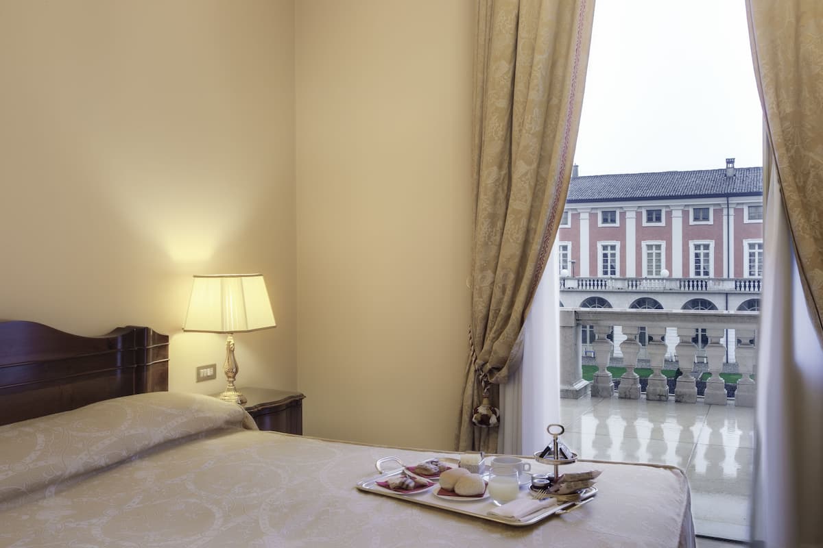 A hotel room with a bed, a tray with breakfast items, a lamp, and a view of a courtyard with buildings.