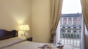 A hotel room with a bed, a tray with breakfast items, a lamp, and a view of a courtyard with buildings.