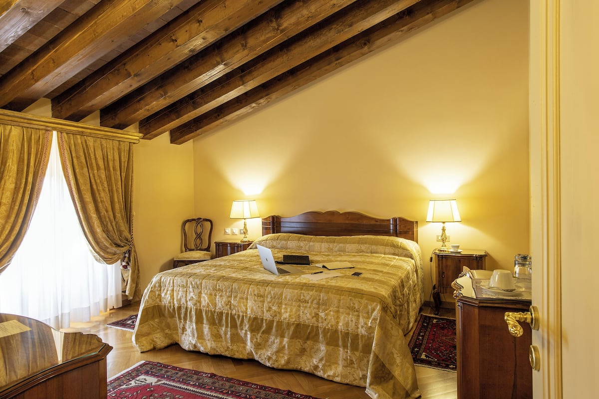 A bedroom with a wooden ceiling, a bed with a laptop, two bedside lamps, and a nightstand with a lamp.