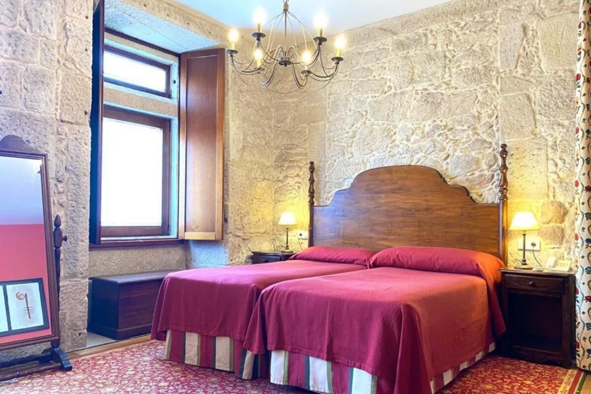 Una habitación con pared de piedra, cama de madera con dos sábanas rojas, un candelabro y una ventana con cortinas.