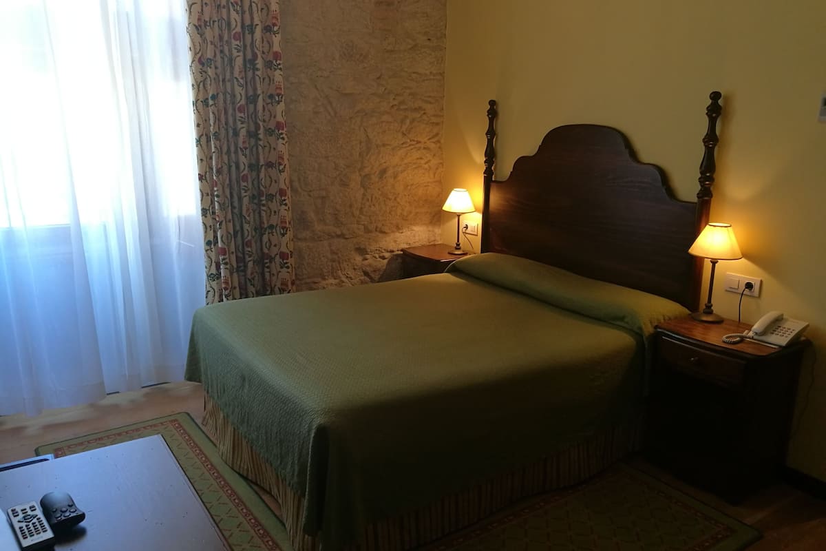 Habitación de hotel con cama, dos lámparas de noche, un teléfono junto a la cama y una mesita con dos controles remotos.