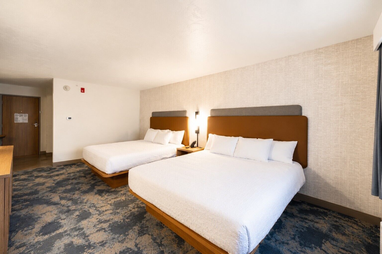 Une chambre d’hôtel avec deux lits, un bureau, une chaise, une porte et un sol recouvert de moquette.