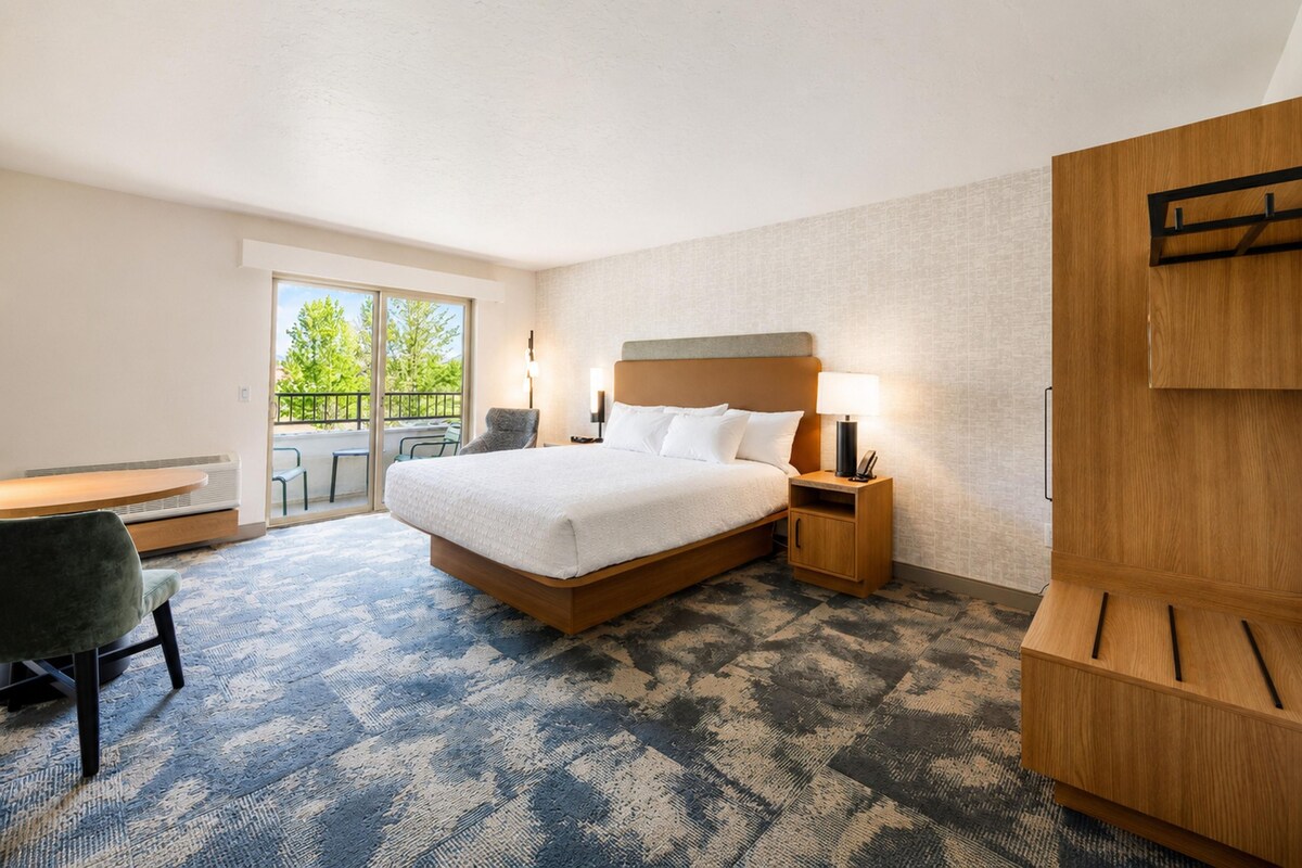 A hotel room with a bed, a desk, a chair, a wardrobe, and a balcony with a view of trees.