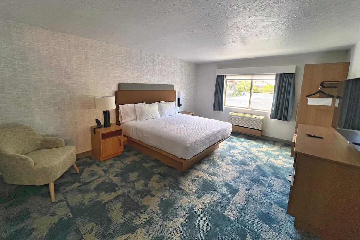 A hotel room with a bed, bedside table, lamp, chair, and a large window.
