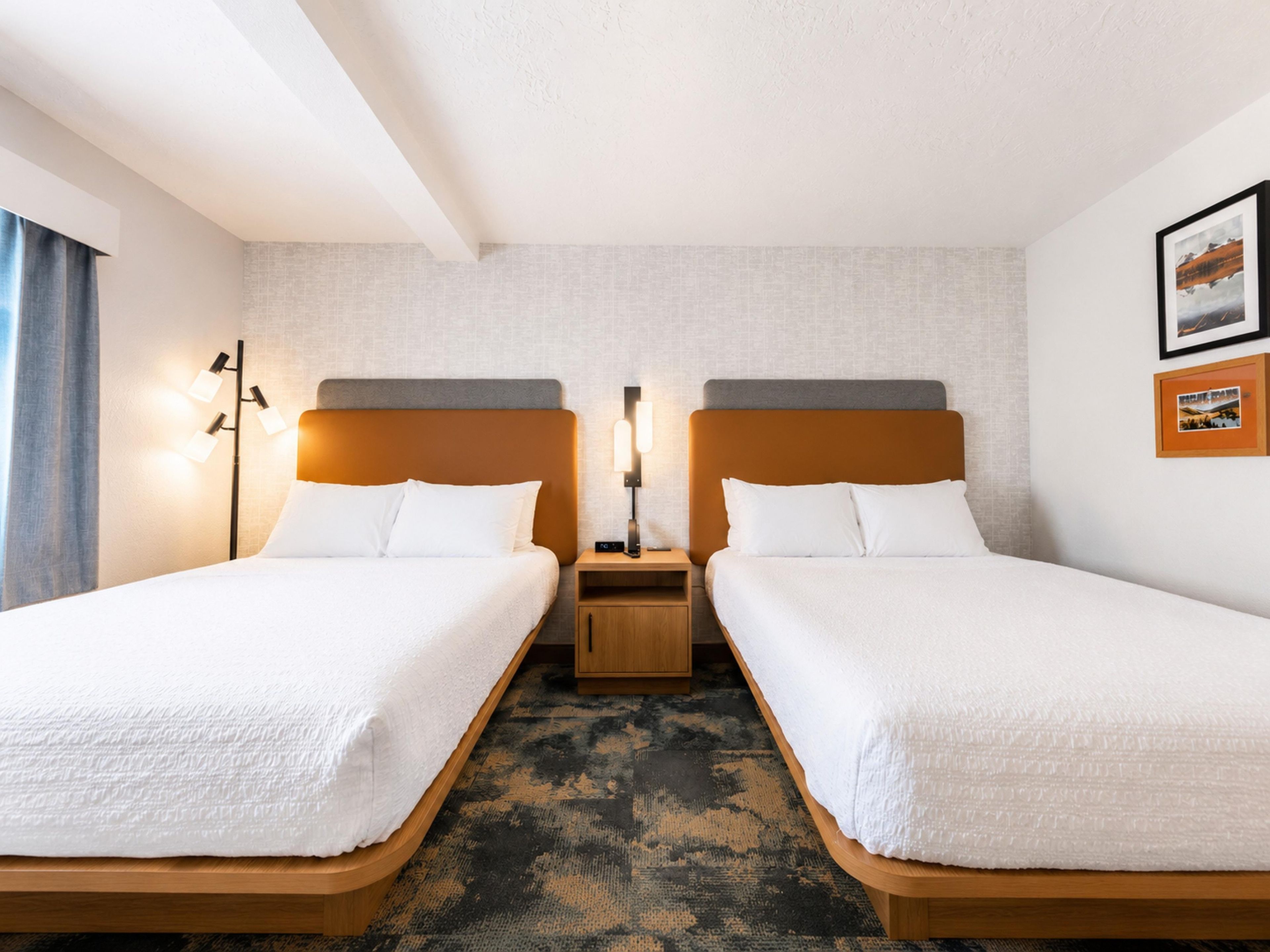 Una habitación de hotel con dos camas individuales, una mesita de noche entre ellas, una lámpara de pared e imagen en un marco colgada en la pared.