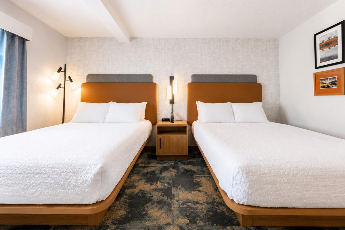 A hotel room with two single beds, a nightstand between them, a wall-mounted lamp, and a framed picture on the wall.