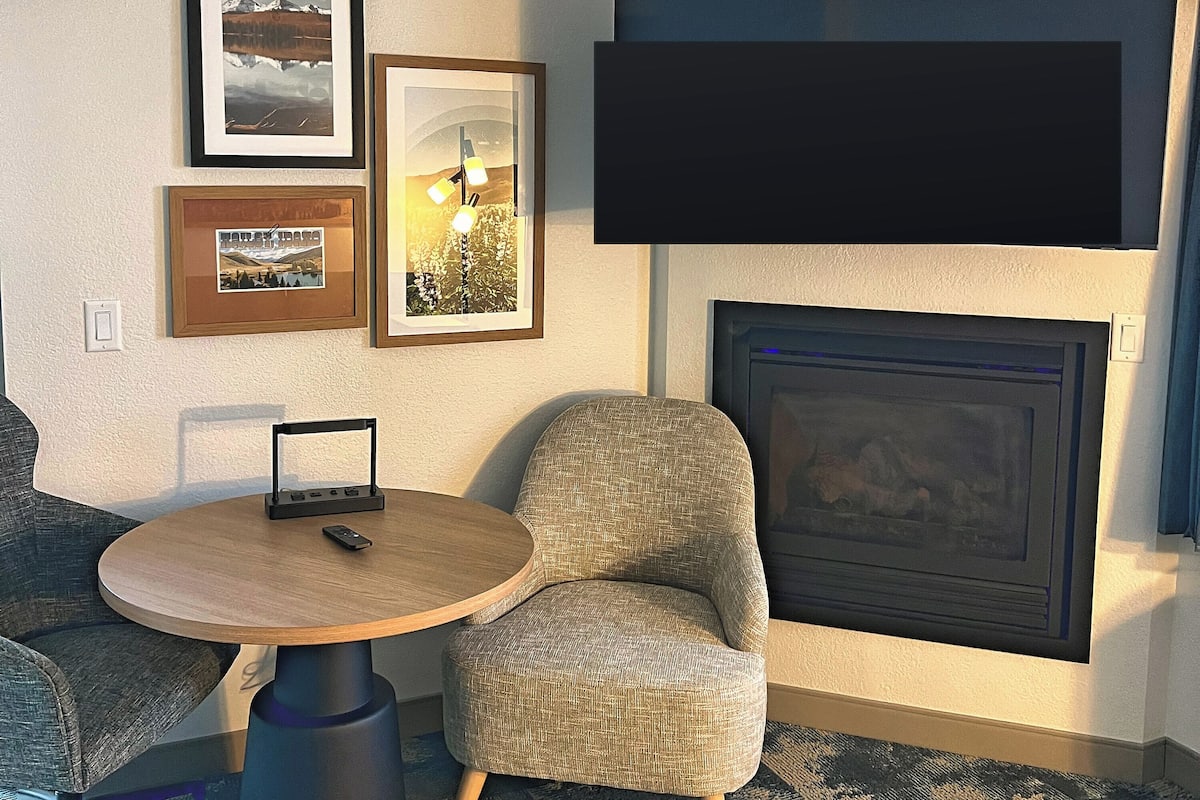 A living room with a fireplace, a round wooden table, a grey armchair, and framed pictures on the wall.