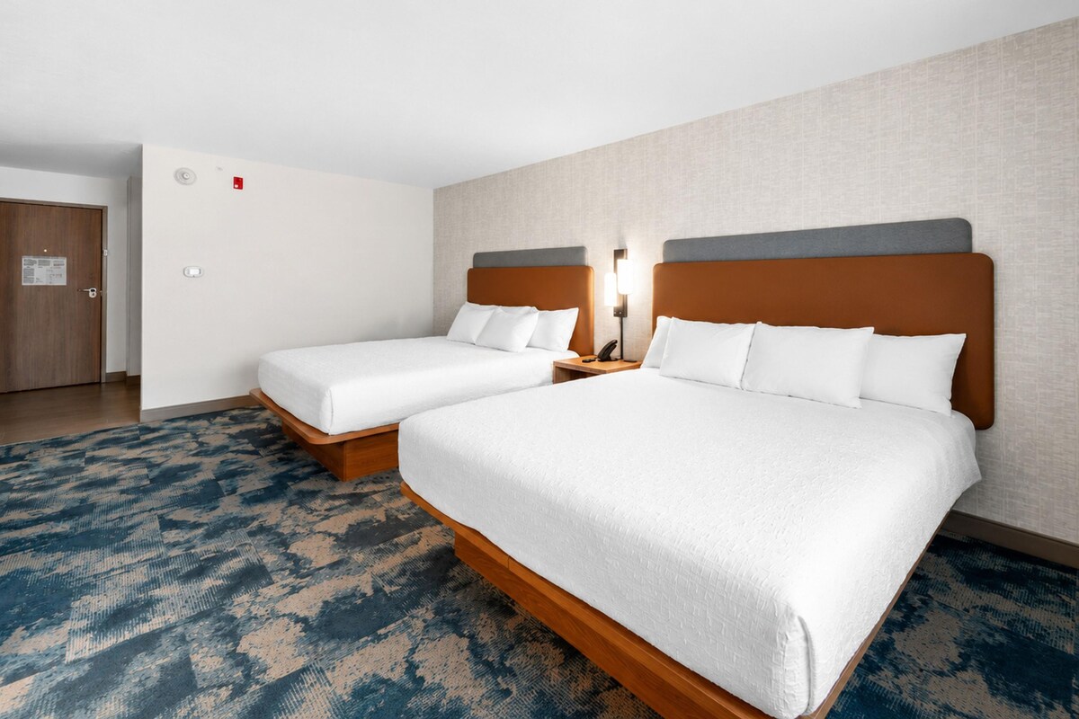 A hotel room with two beds, a wooden floor, a patterned carpet, and a plain wall.