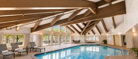 Uma piscina interior com chão em mosaico, espreguiçadeiras e teto alto com vigas aparentes.