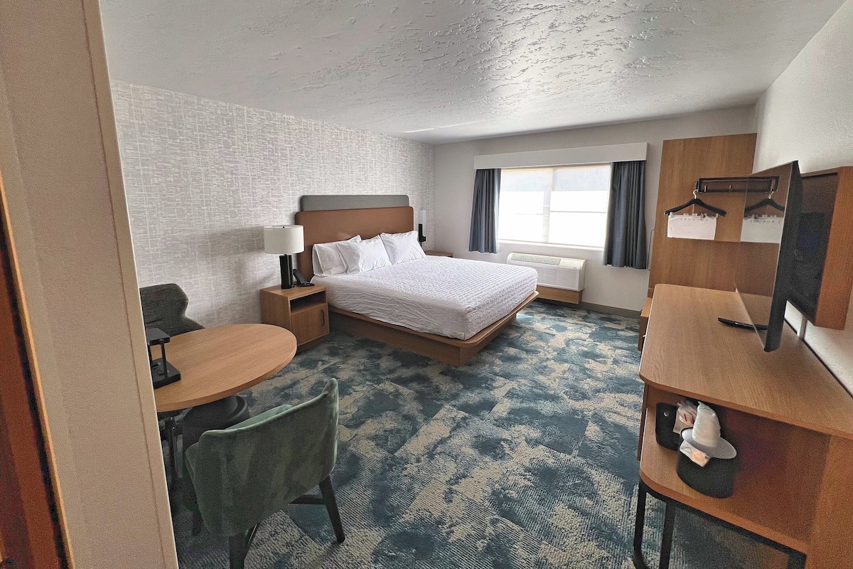 A hotel room with a bed, bedside table, lamp, chair, desk, and a window with curtains.