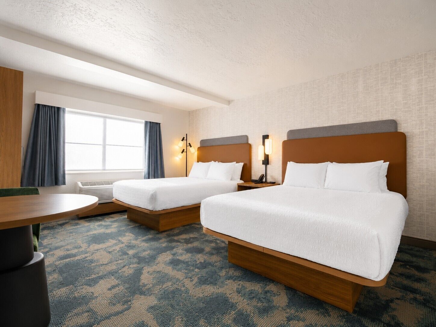 A hotel room with two beds, a desk, a chair, a window with curtains, and a wall-mounted lamp.
