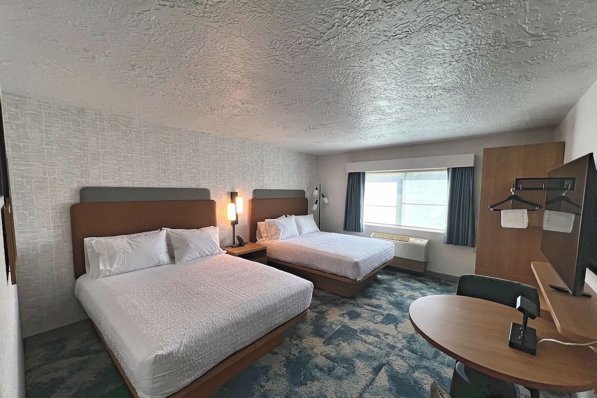 A hotel room with two beds, a desk, a chair, a window with curtains, and a wall-mounted coat rack.