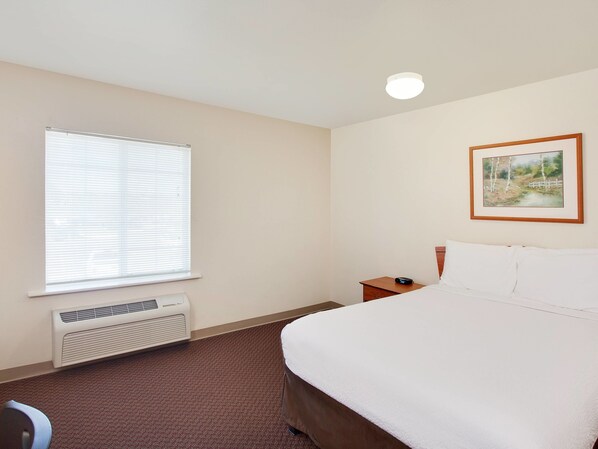 A hotel room with a large bed, a bedside table, a nightstand, a window with blinds, a wall-mounted air conditioner, and a framed picture on the wall.