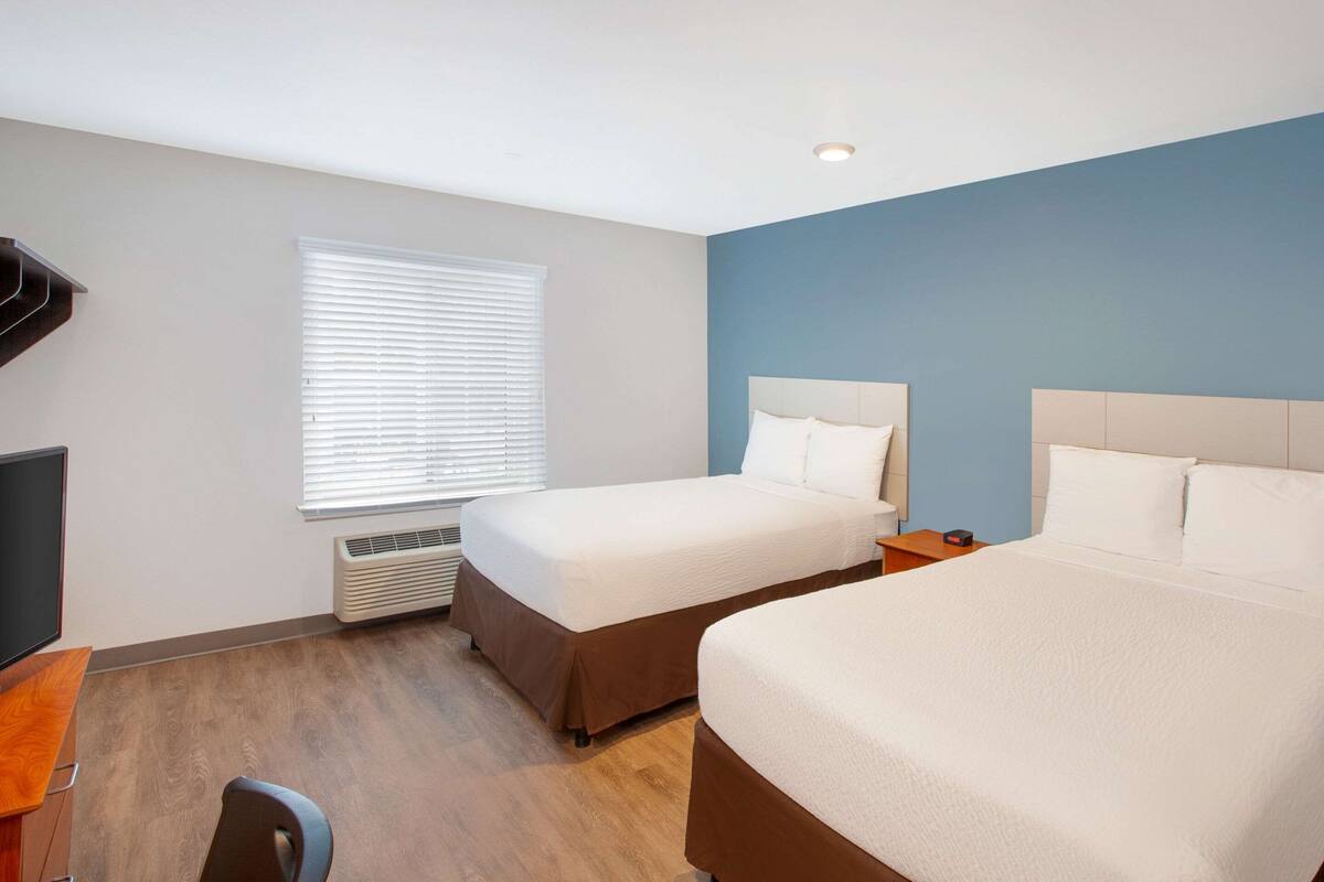 Une chambre d’hôtel avec deux lits, un bureau, une chaise, une télévision et une fenêtre avec des stores.
