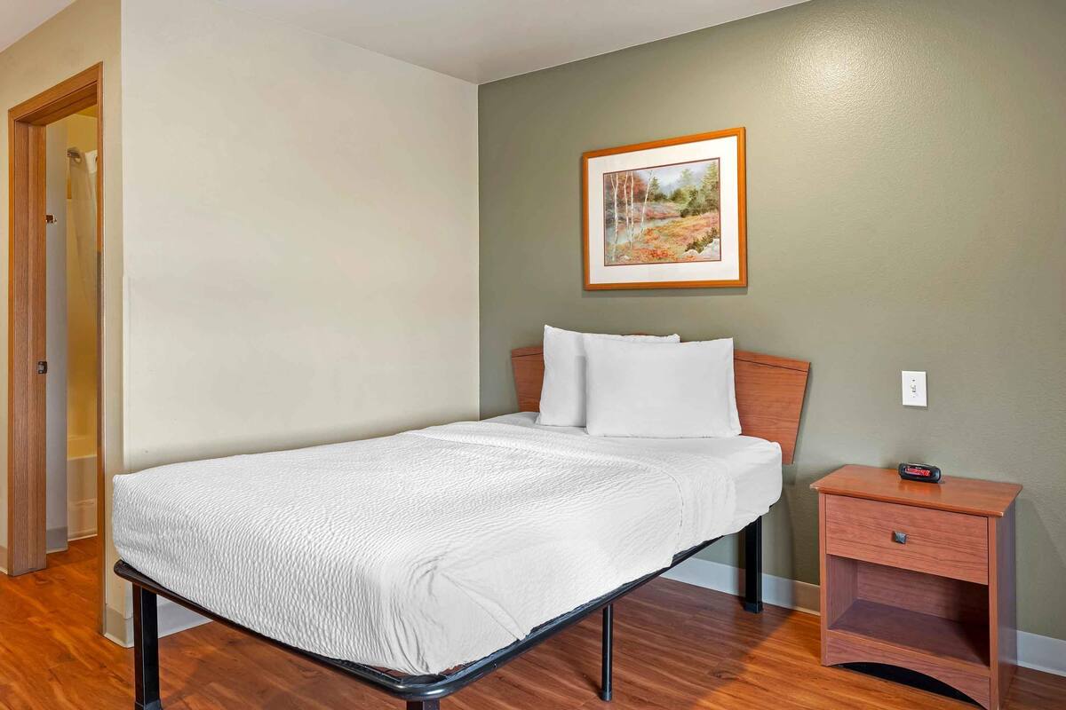 Una cama individual con colchón y almohadas blancas, una mesita de noche con cajón, un cuadro en la pared y una puerta de madera.