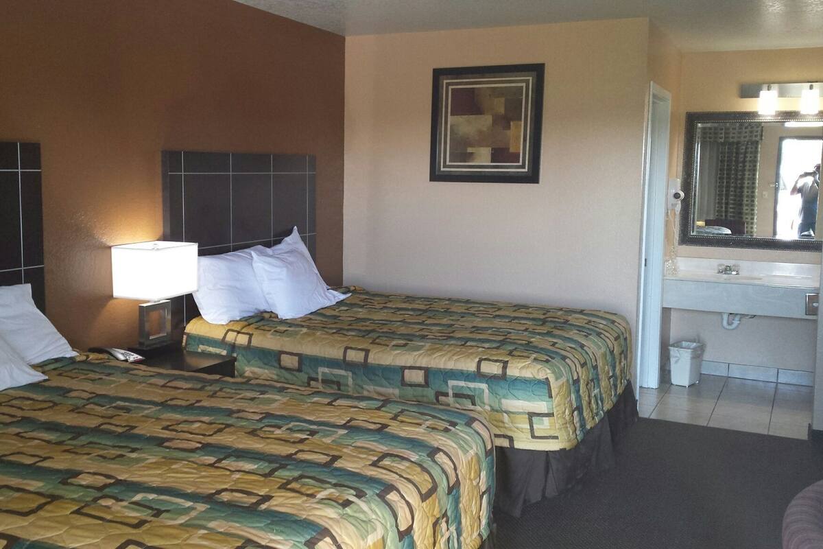 A hotel room with two beds, a nightstand with a lamp, a bathroom with a sink and mirror, and a framed artwork on the wall.