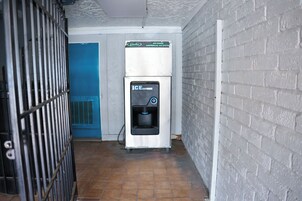 Un dispensador de hielo comercial en un pasillo estrecho con una puerta azul y una pared de ladrillo blanco.