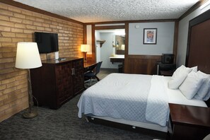 A hotel room with a brick wall, a flat-screen TV, a desk with a lamp, a bed with white bedding, and a wooden headboard.
