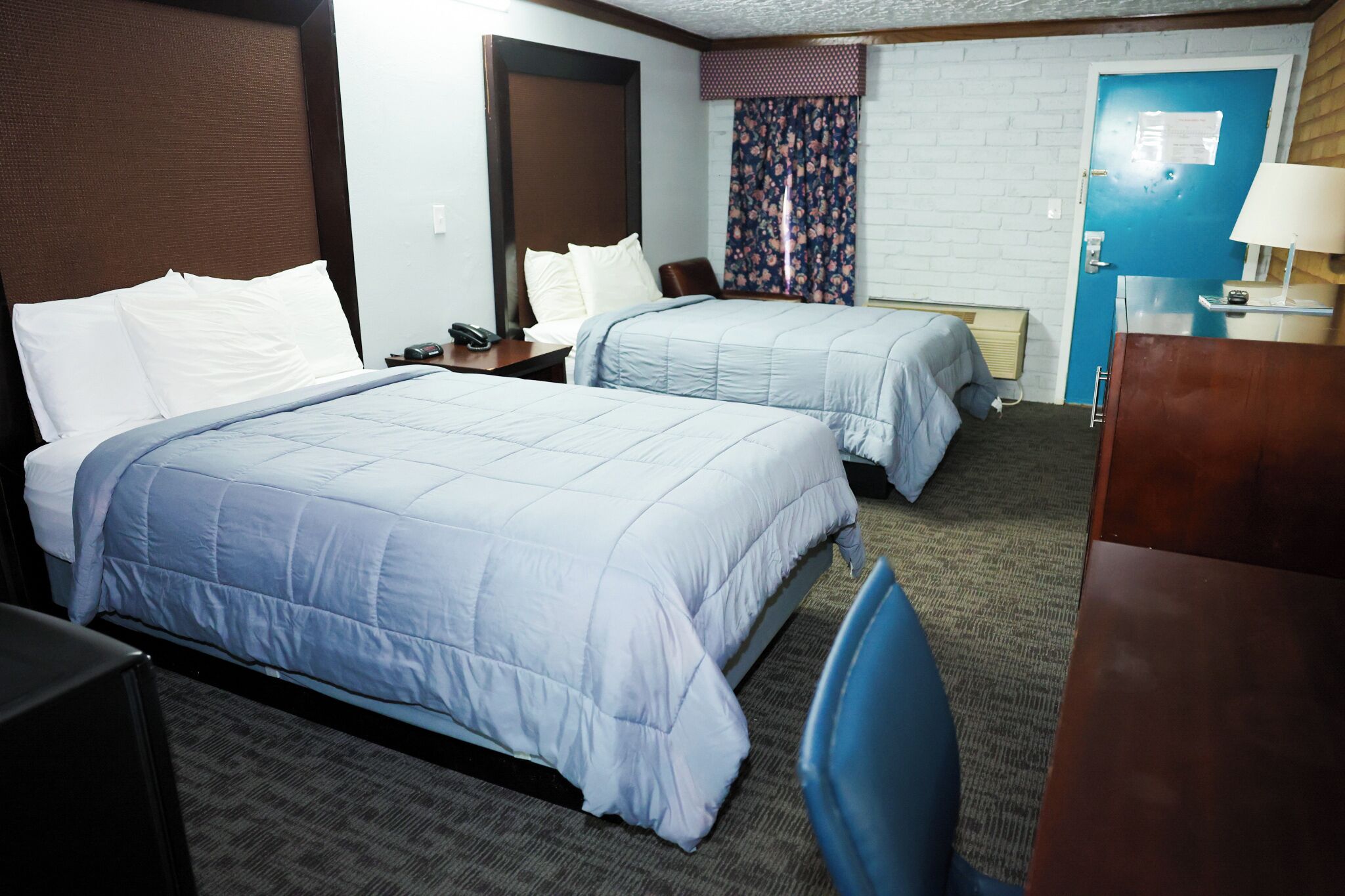 A hotel room with two beds, a desk, a chair, a lamp, and a door with a sign.