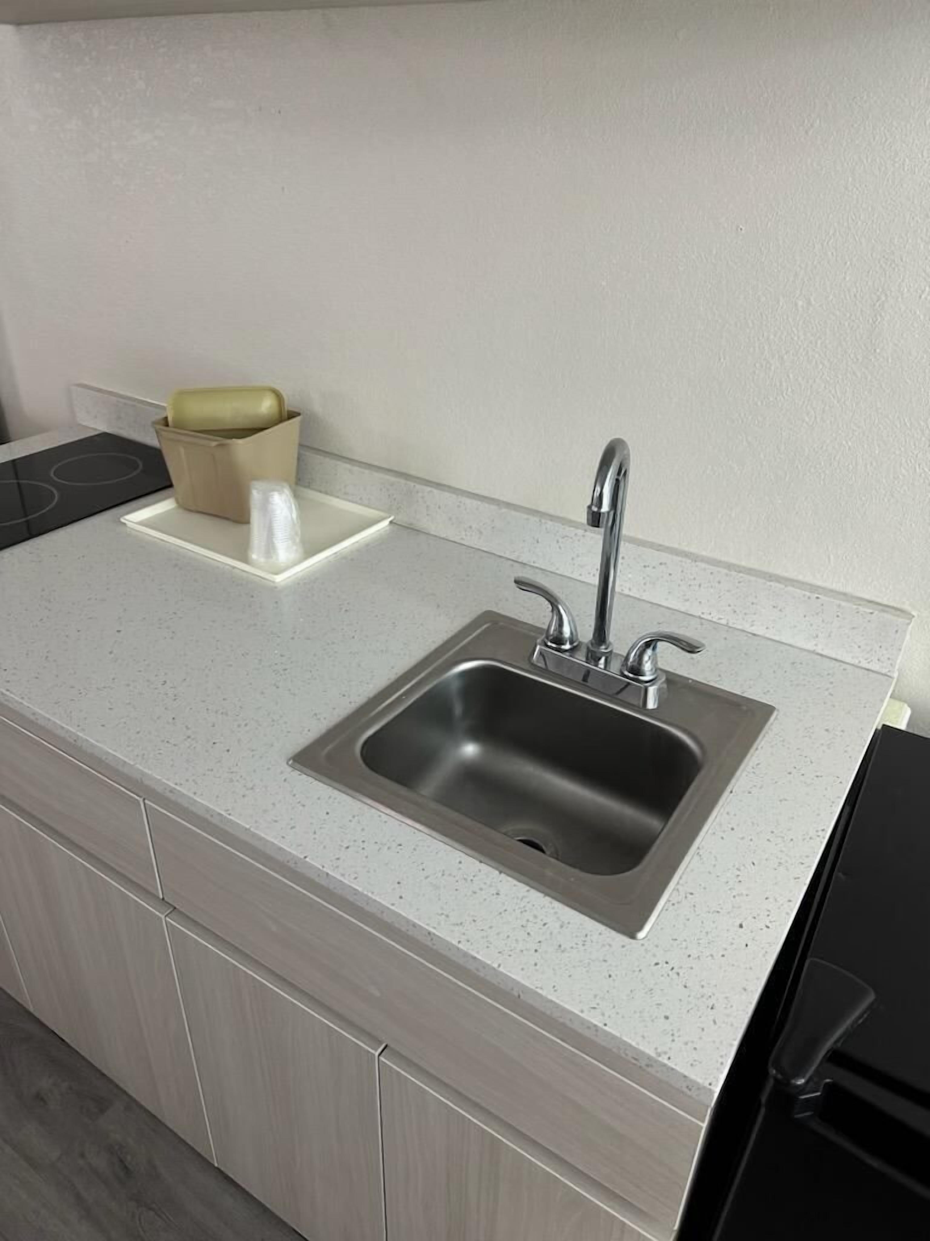 A kitchen with a stainless steel sink, a beige trash bin, and a white countertop.