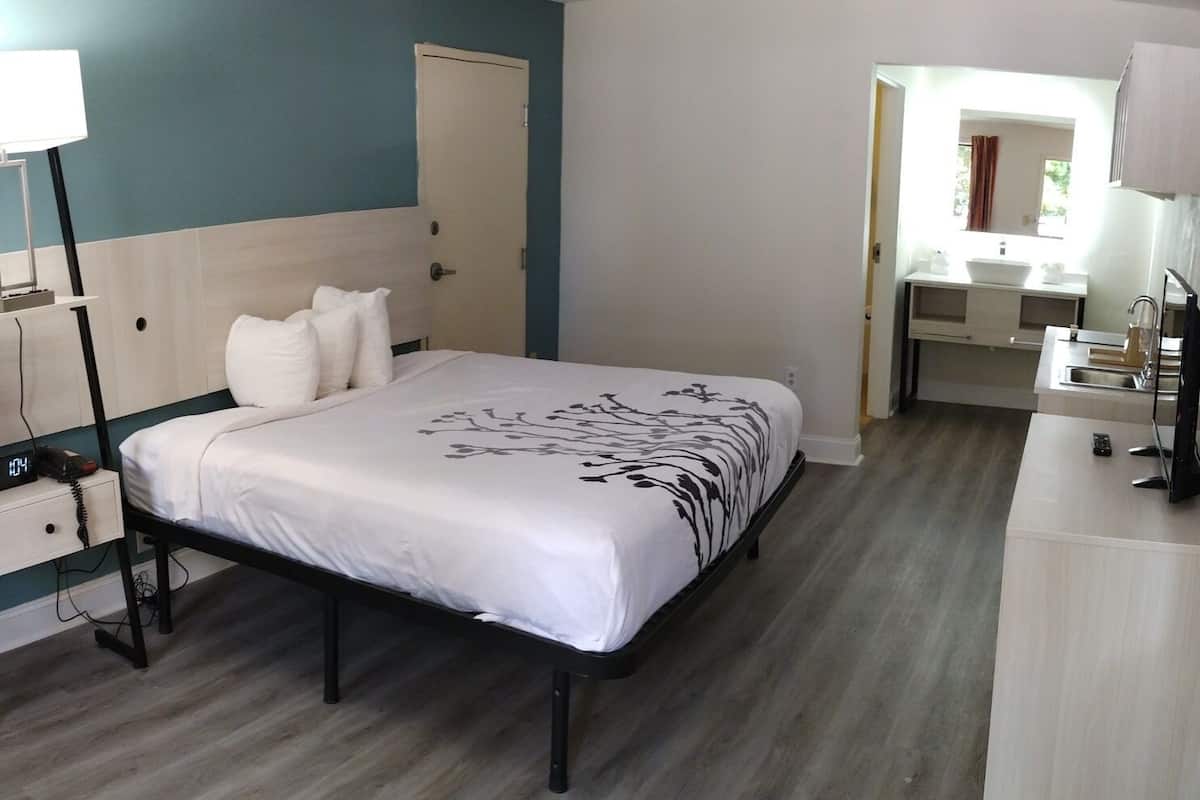 Habitación de hotel con cama, mesita de noche, lámpara y baño visible a través de una puerta abierta.