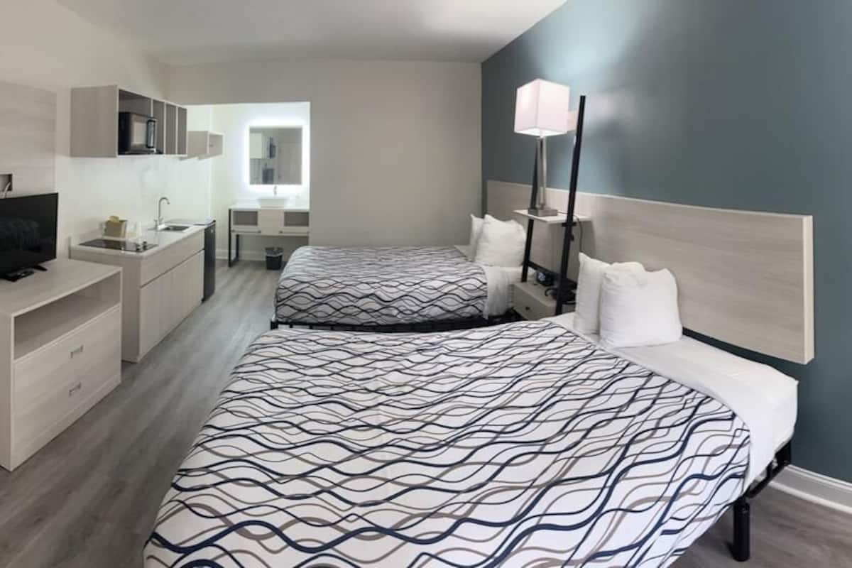 Una habitación de hotel moderna con dos camas, una kitchenette pequeña, un espejo y una lámpara de noche.