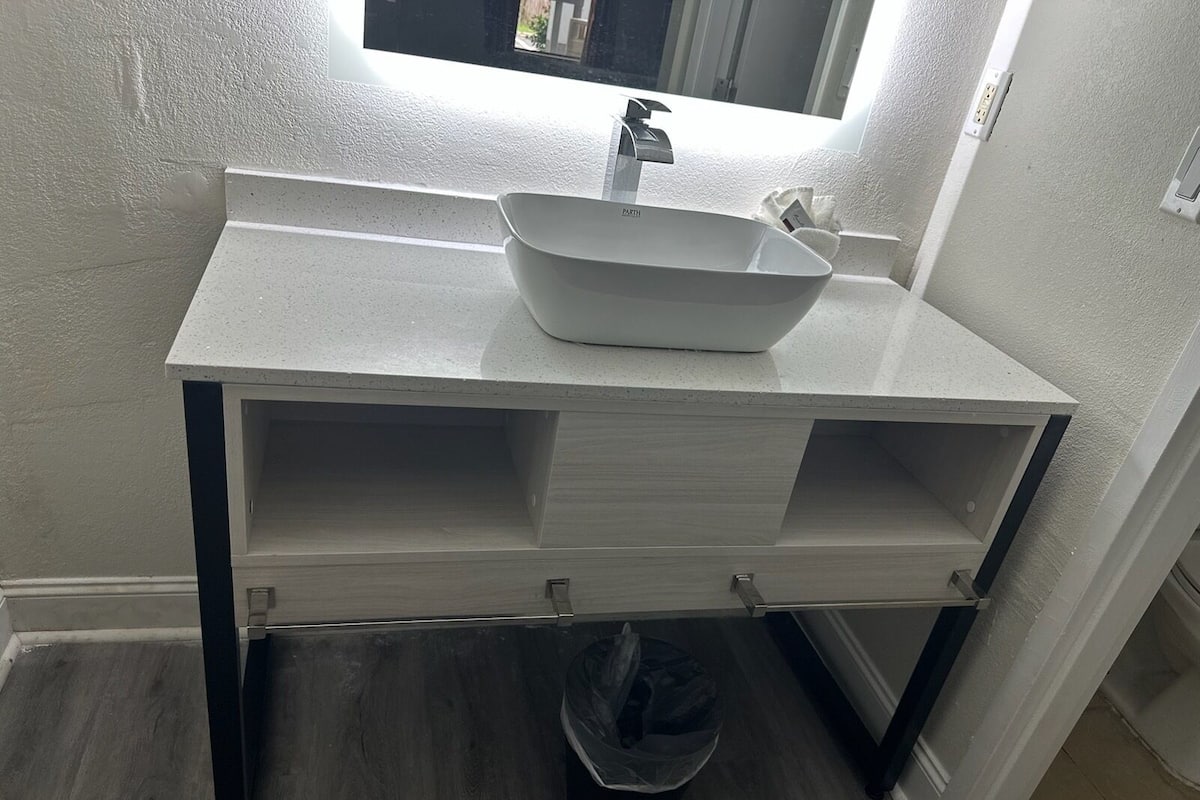 Un baño con un lavamanos blanco, un espejo encima y un cubo de basura debajo.