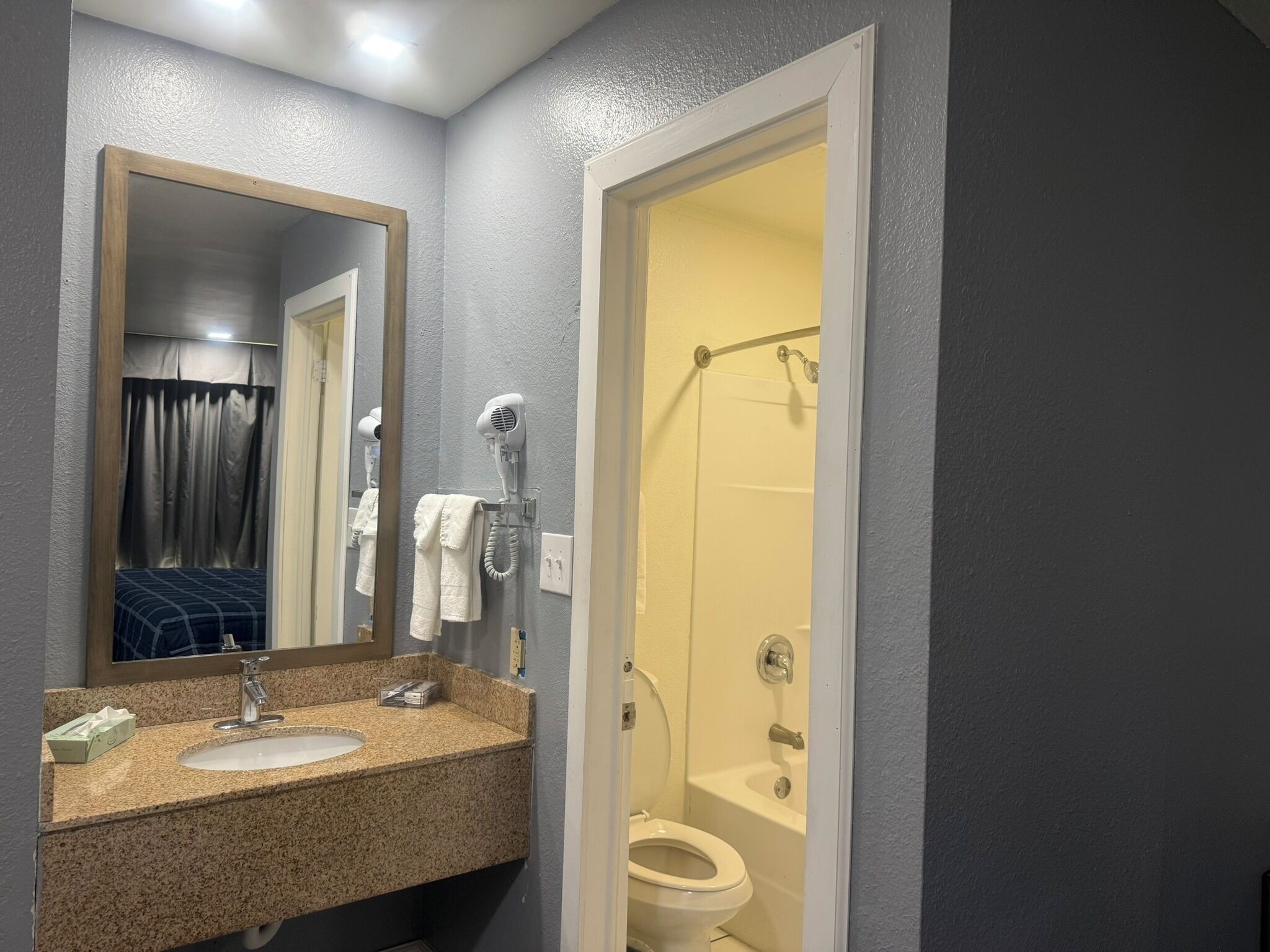 A bathroom with a granite countertop sink, a mirror, a towel rack, a hairdryer, and a shower stall.
