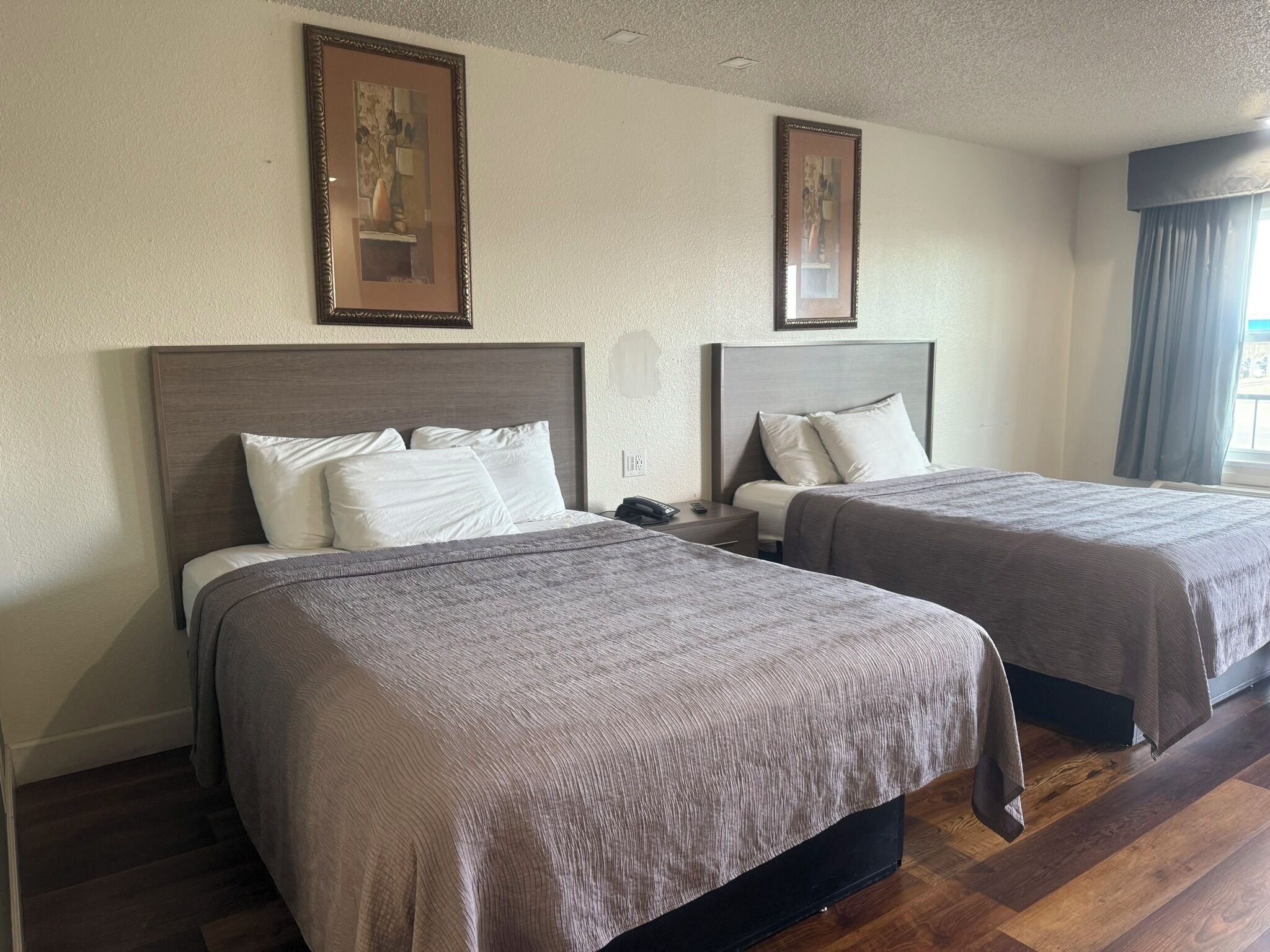A hotel room with two beds, each with a wooden headboard and white bedding. There are two framed pictures on the walls, and a window with curtains.