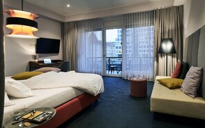 A hotel room with a bed, a desk with a laptop, a TV, a sofa, and a view of buildings through the window. - Hotel Neuer Fritz Berlin (Berlin)