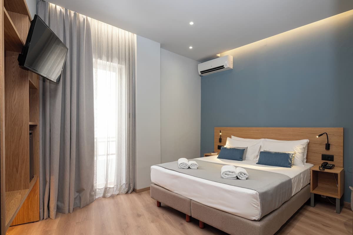 Una habitación de hotel moderna con una cama grande, un televisor de pantalla plana montado en la pared, un equipo de aire acondicionado y una mesita de noche de madera con lámpara.