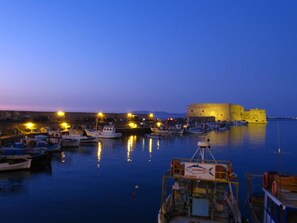 A harbor at dusk with boats docked, a fortified building, and a clear sky. - Athinaikon (Heraklion)