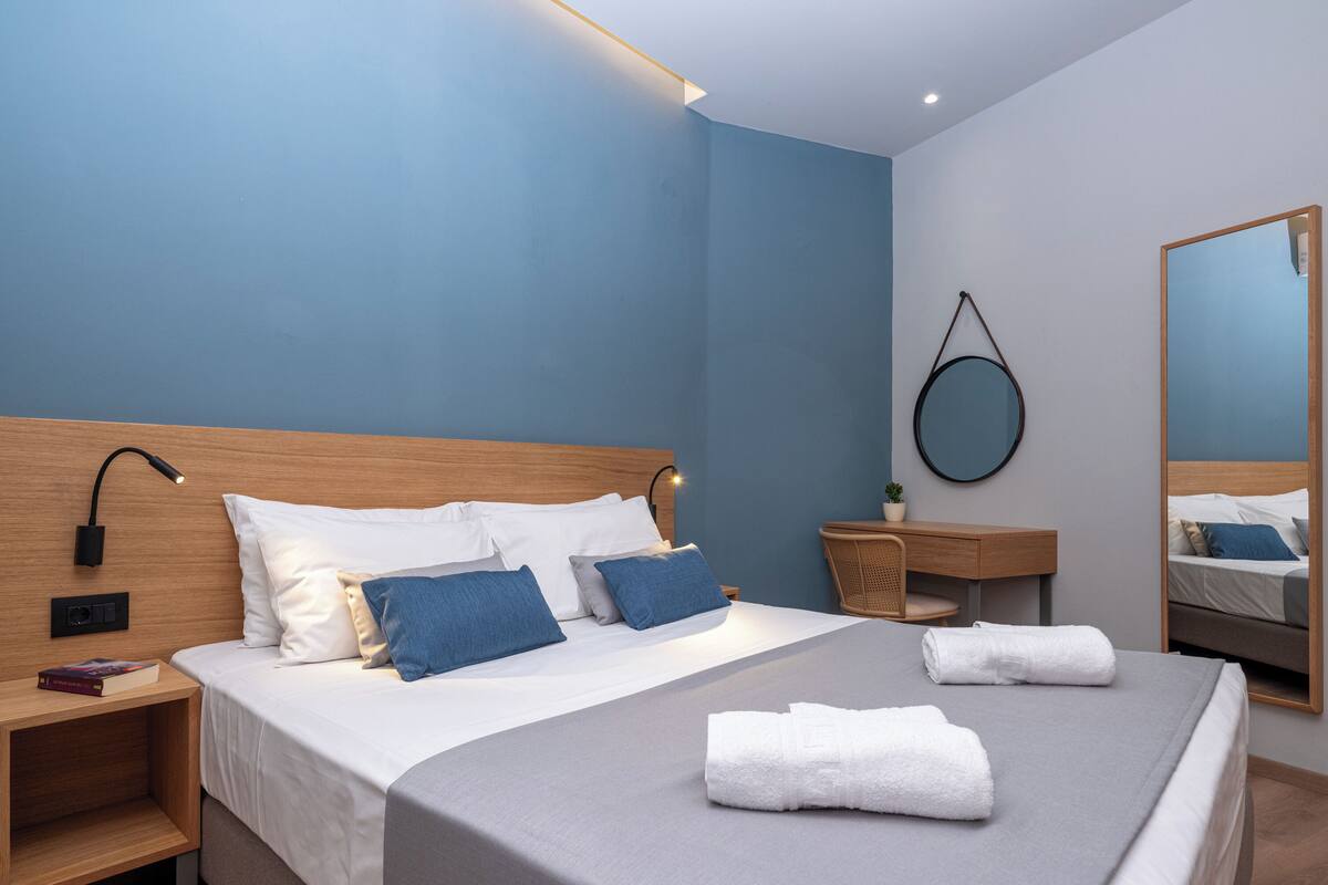 Una habitación de hotel moderna con un cabecero de madera, una cama grande con ropa de cama blanca y azul, una mesita de noche con una lámpara, un espejo redondo y una pequeña planta.