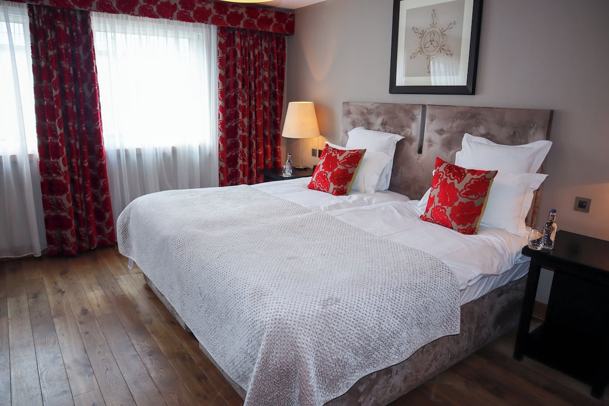 Una cama bien tendida con sábanas blancas y almohadas rojas, una lámpara de noche y un cuadro en la pared.