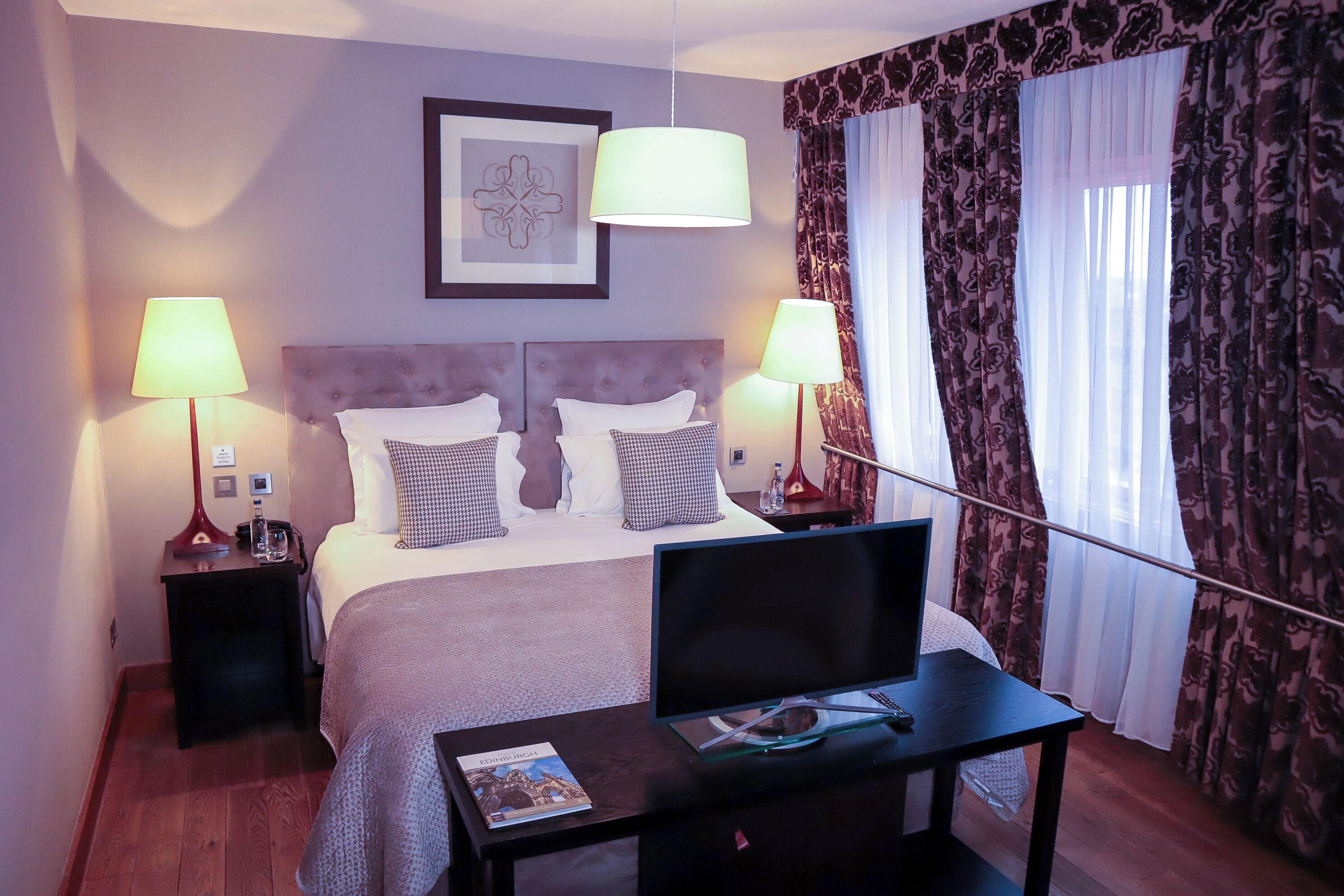 A hotel room with a bed, two bedside lamps, a TV on a stand, and a framed picture on the wall.