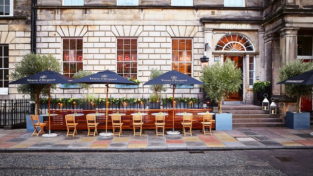 An outdoor seating area with wooden chairs and tables, umbrellas, and potted plants. - Tigerlily (Edinburgh)