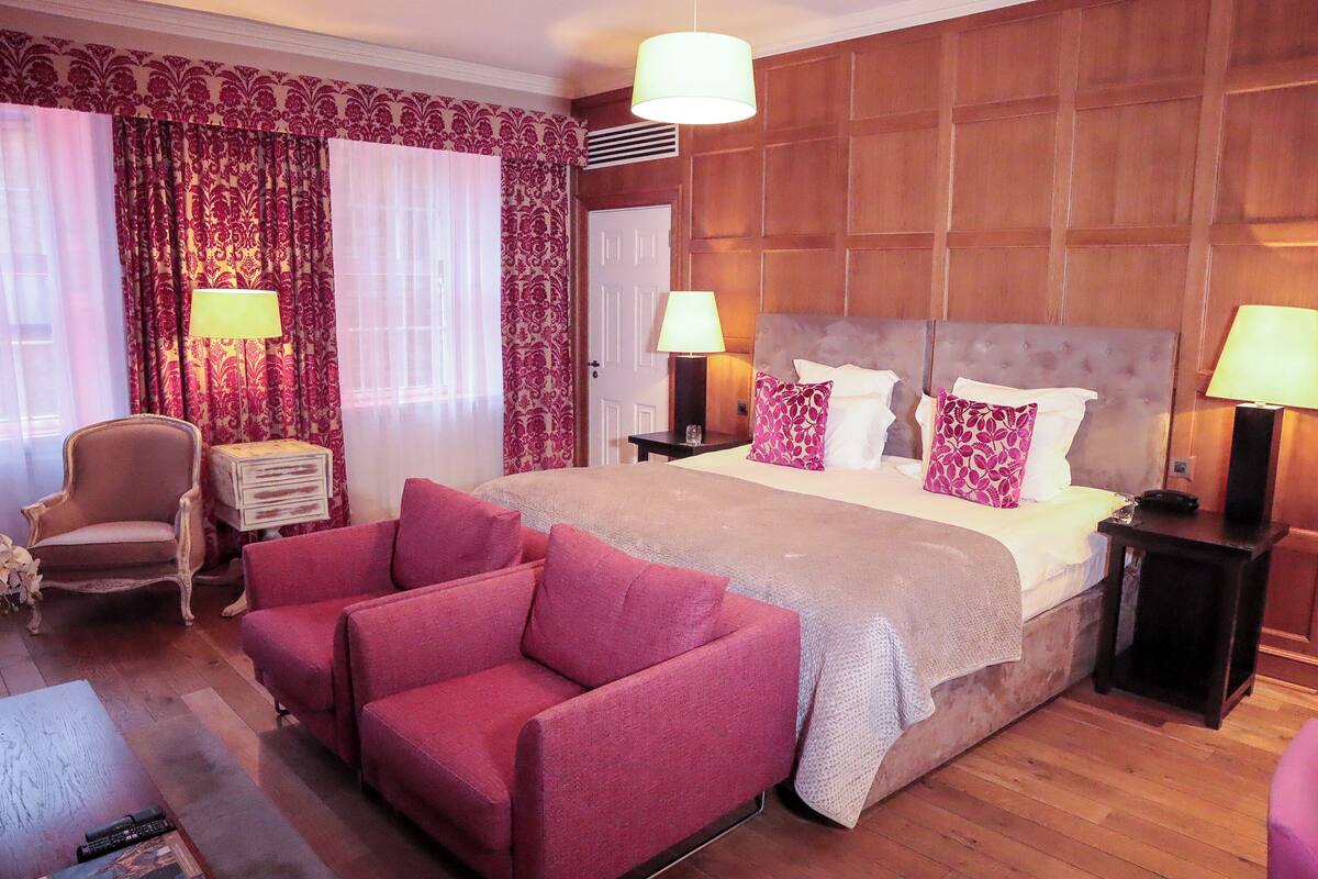 Una habitación de hotel con una cama grande, dos mesitas de noche con lámparas, un sillón rojo, un cabecero de madera y una cortina estampada.
