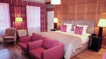 Quarto de hotel com uma cama grande, duas mesas de cabeceira com abajures, uma poltrona vermelha, um painel de cabeceira de madeira e cortina estampada.