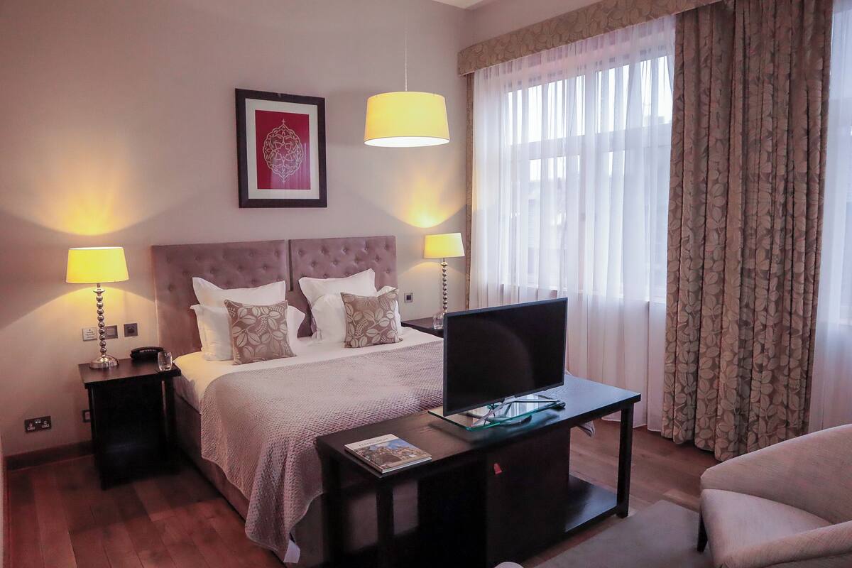 Habitación de hotel con cama, dos lámparas de noche, un televisor de pantalla plana sobre una mesita de vidrio, un cuadro en la pared y una ventana con cortinas.
