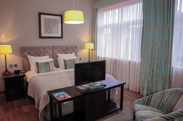Habitación de hotel con cama, dos lámparas de noche, televisor sobre un mueble bajo, una mesita con una revista y una ventana con cortinas.