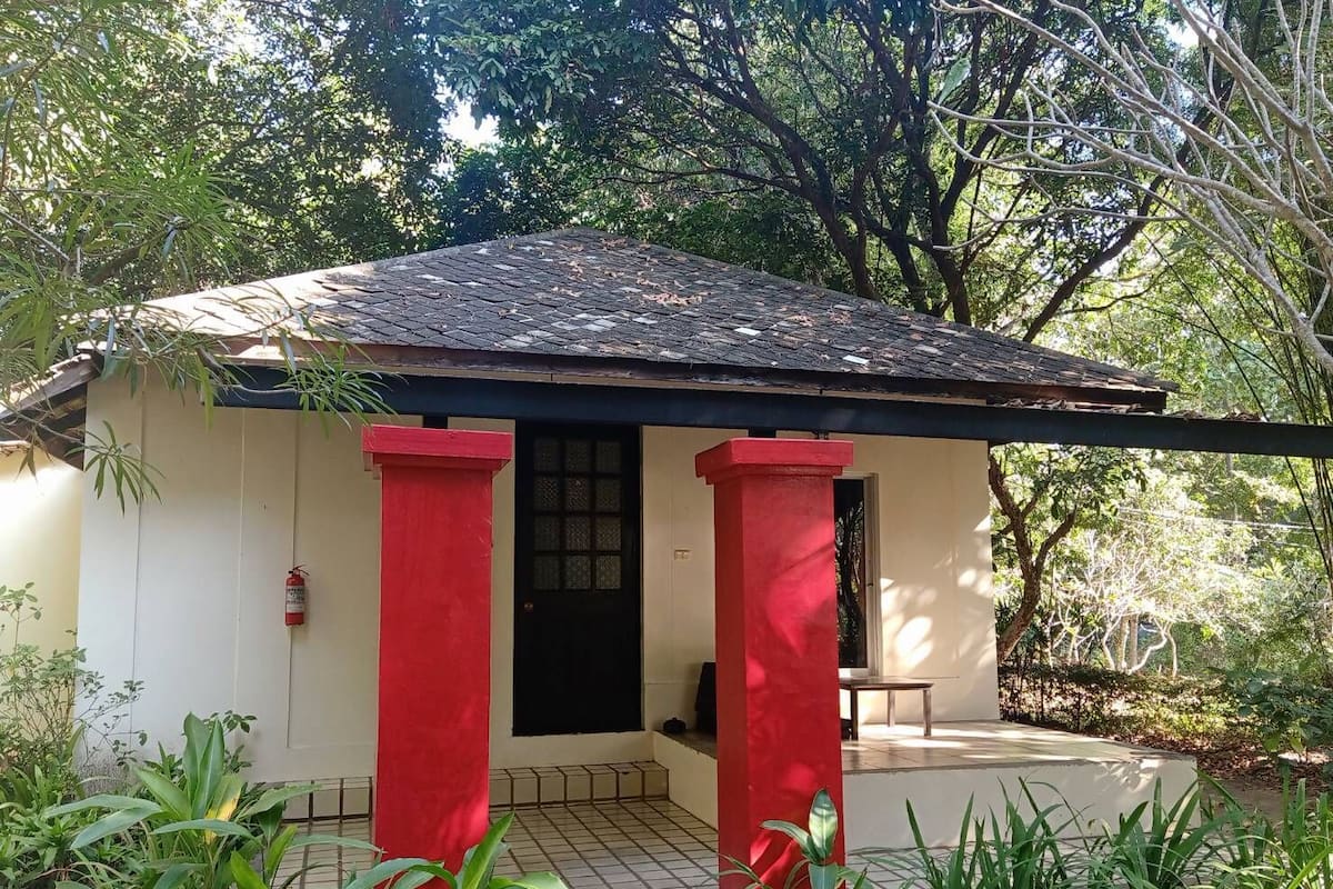 A small, single-story building with a tiled roof and a black door. It has a red column on each side and is surrounded by greenery.