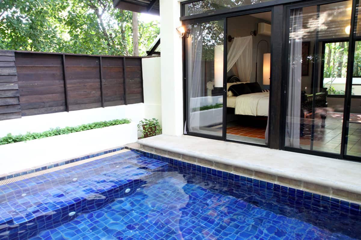 A swimming pool with blue mosaic tiles, a wooden fence, and a glass door leading to a bedroom with a bed and a nightstand.