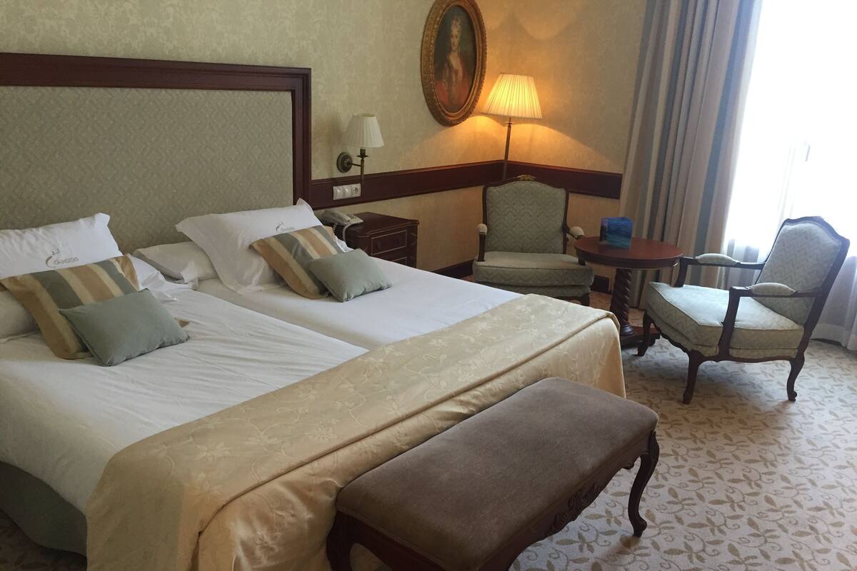 A hotel room with two beds, a bench, a chair, a lamp, and a painting on the wall.