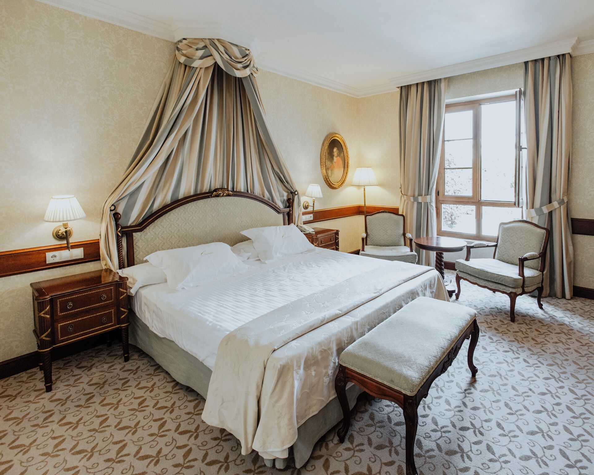 A hotel room with a large bed, a bedside table, a chair, a small table, and a window with curtains.