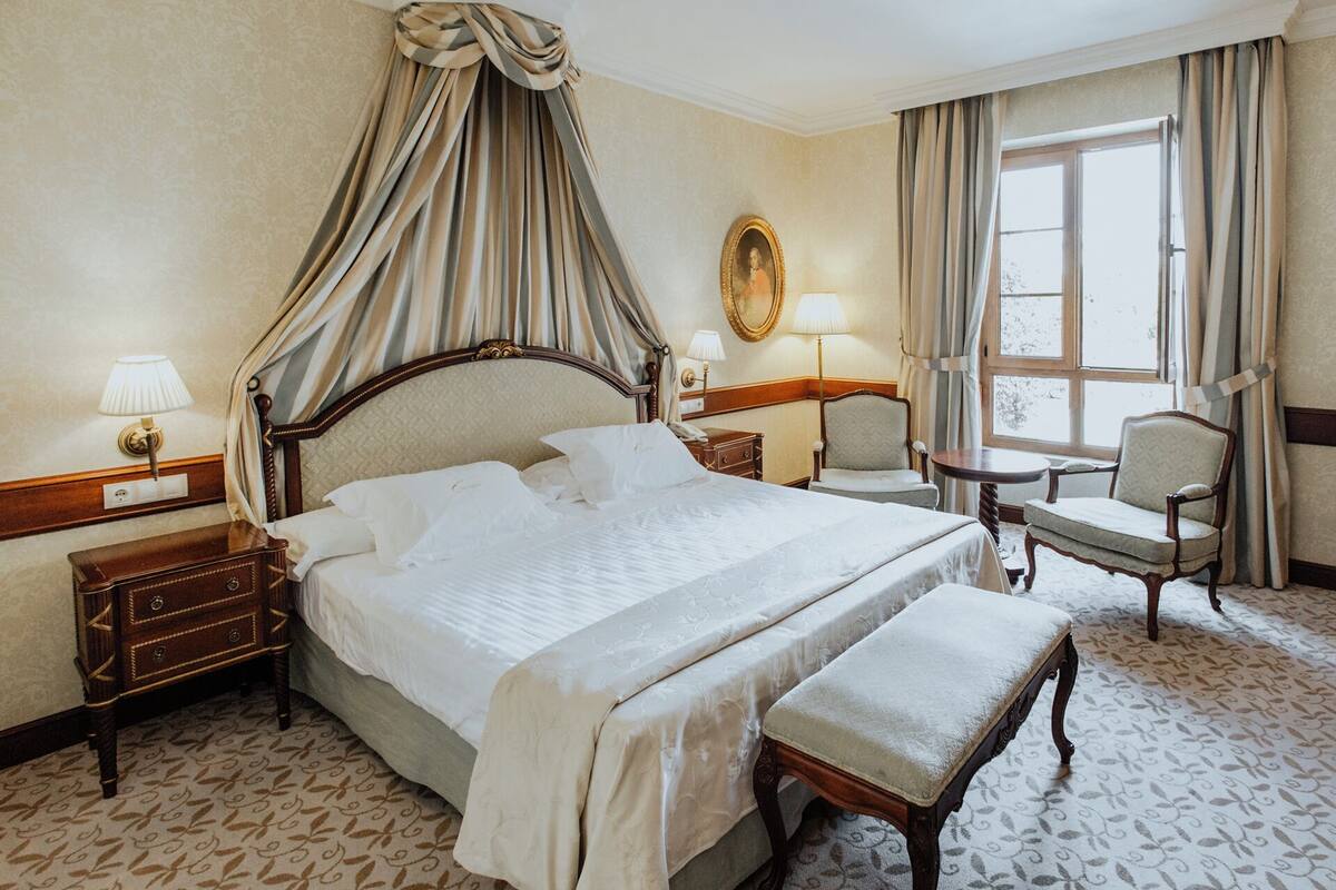 A hotel room with a large bed, a bedside table, a chair, a small table, and a window with curtains.