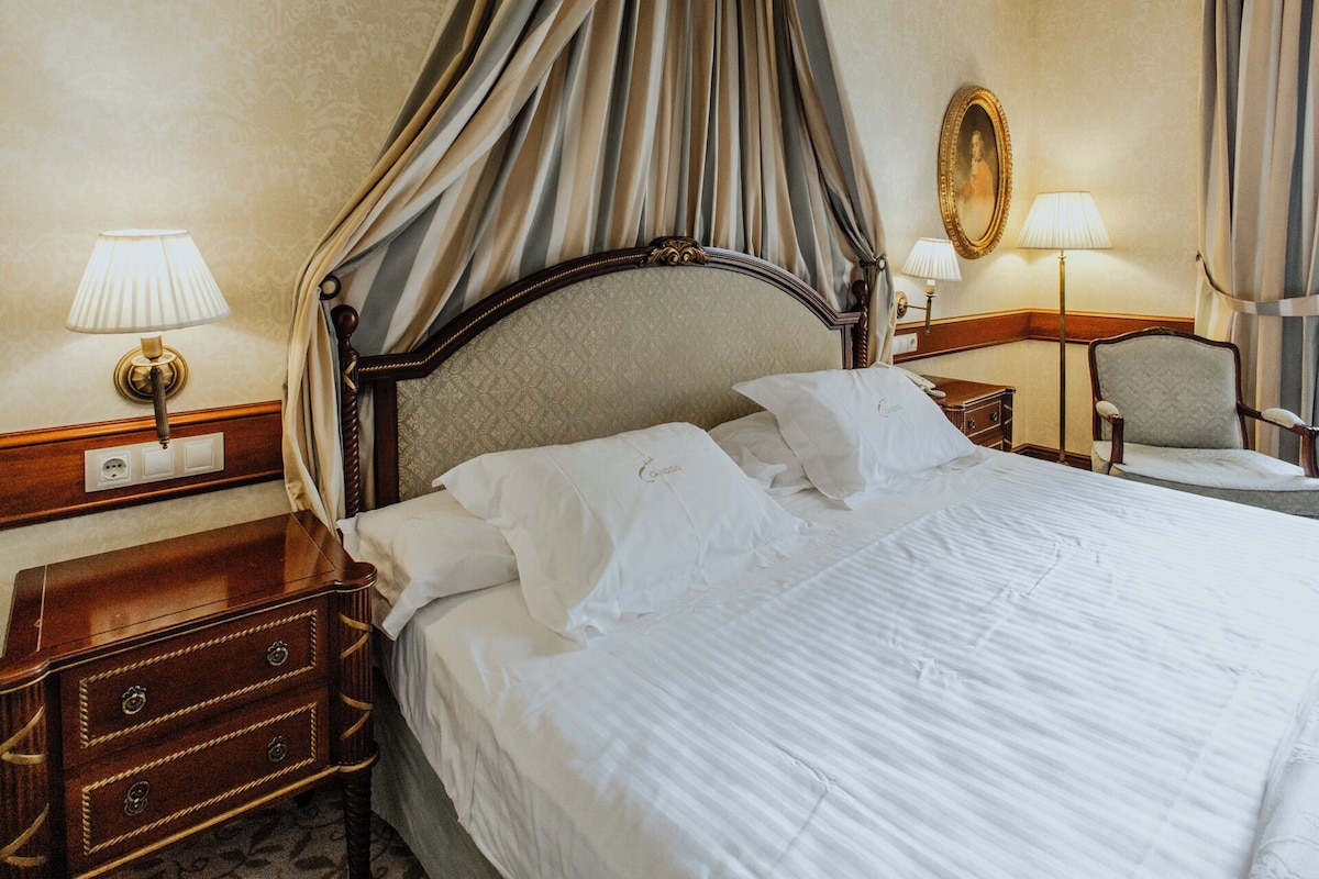 A hotel room with a four-poster bed, a nightstand with a lamp, a chair, and a mirror on the wall.