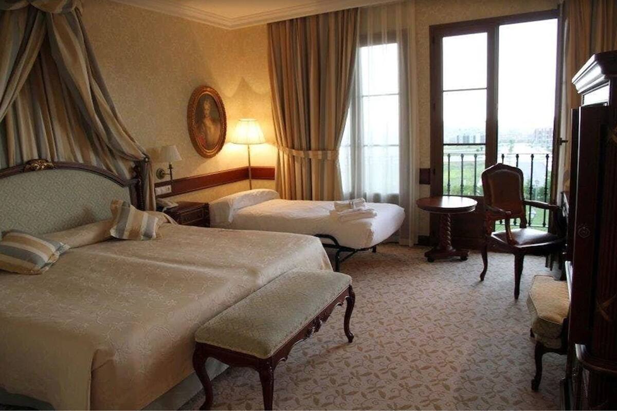 A hotel room with two beds, a sitting area with a chair, a lamp, and a painting on the wall.